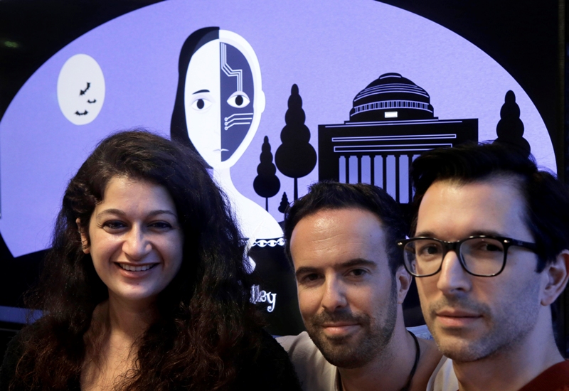 In this Tuesday, Oct. 24, 2017 photo creators of a fiction-writing 'chatbot,' from the left, Massachusetts Institute of Technology postdoctoral associate Pinar Yanardag, of Istanbul, Turkey, MIT research scientist Manuel Cebrian, of Madrid, Spain, and MIT associate professor Iyad Rahwan, of Aleppo, Syria, sit for a photograph in front of a graphic from the home page of the site called "Shelley." (AP Photo/Steven Senne)