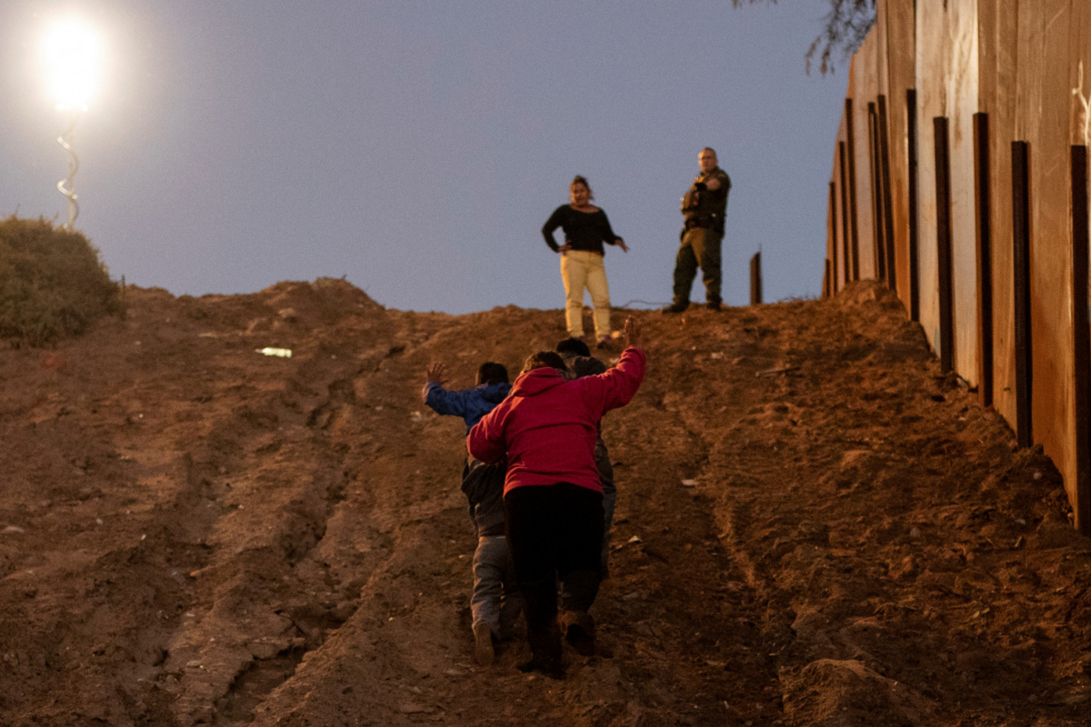 Migrants from Honduras, part of a caravan of thousands from Central America trying to reach the United States, put their hands in the air as they surrender to a U.S. Customs and Border Protection official in San Diego County, photographed from Tijuana, Mexico, on Dec. 3, 2018. (Alkis Konstantinidis/Reuters)