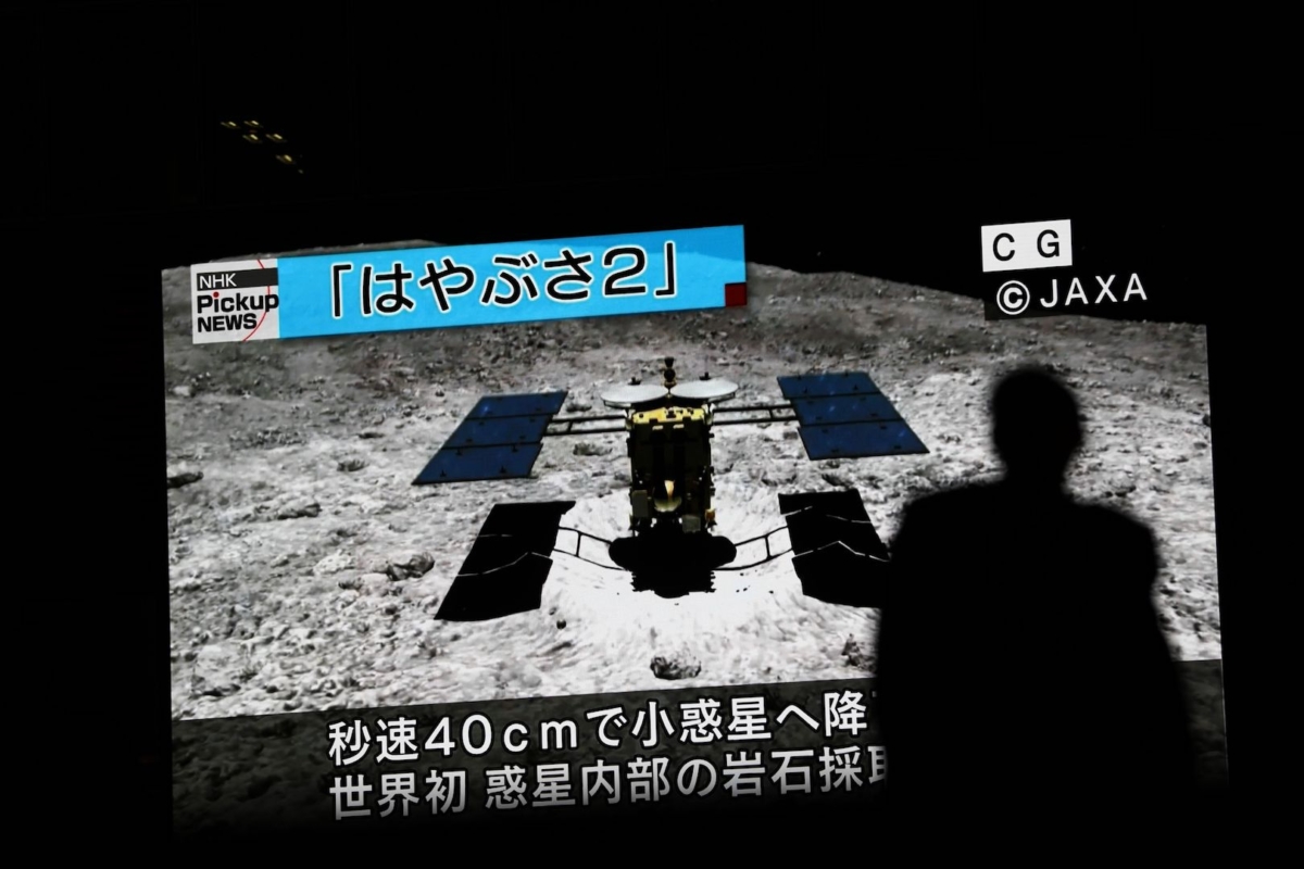 In this picture taken on July 10, 2019 a man walks past a screen displaying a computer-generated image of the Hayabusa2 probe, during a news broadcast at Akihabara district in Tokyo. (Photo by Behrouz Mehri/AFP via Getty Images)
