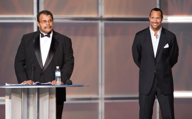 In this 2008 photo provided by WWE, Inc., Rocky "Soul Man" Johnson, left, speaks at his WWE Hall of Fame induction ceremony, as his son, Dwayne “The Rock” Johnson watches. (WWE, Inc. via AP)