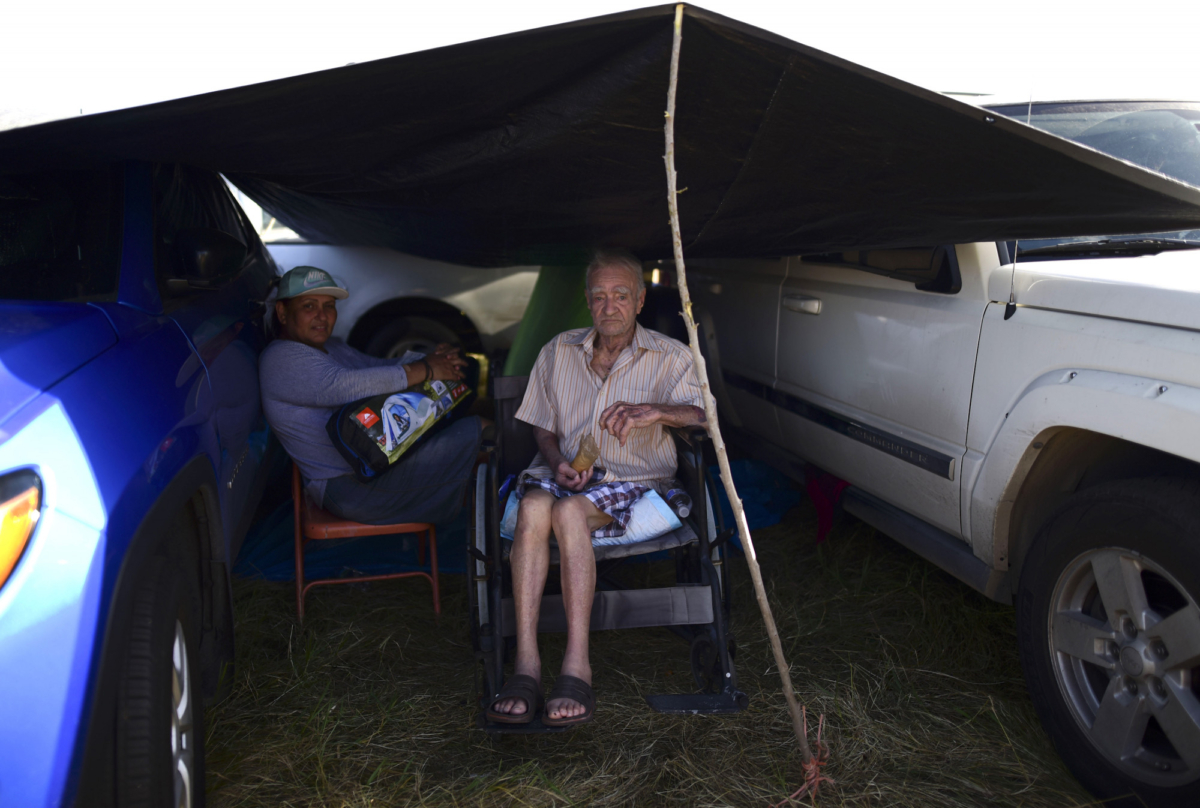 Residents from the Indios neighborhood of Guayanilla, Puerto Rico, Milagros Figueroa and Ruben Fantausi, sit under a tarp between vehicles parked on a private hay farm where locals affected by earthquakes have set up shelter amid aftershocks in Guayanilla, Puerto Rico, on Jan. 10, 2020. (Carlos Giusti/AP Photo)