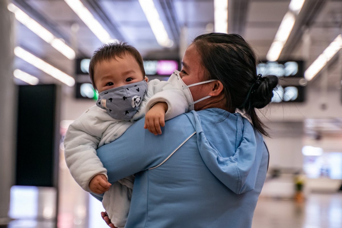 A woman carries a baby wearing a protective mask