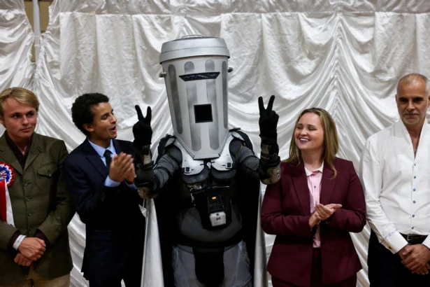 Count Binface gestures while standing with other candidates after Reform UK party leader Nigel Farage was declared the winner for the Clacton by-election, triggered by the resignation of Farage, in Essex, Britain, on Aug. 14, 2026. (Jack Taylor/Reuters)